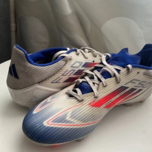 Adidas F50 fotbollsskor blå/röd - Säljer ett par Adidas F50 fotbollsskor i vitt, blått och rött med sportig design och snörning. Skorna har en lätt ovandel i syntetmaterial och klassisk Adidas-logga på hälen. Perfekta för dig som vill ha snabbhet och stil på planen.