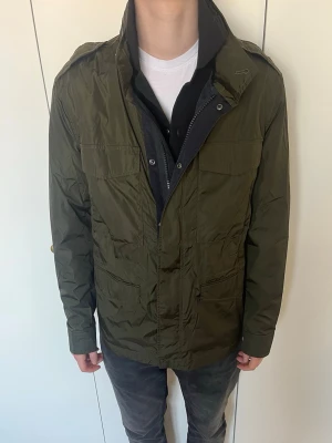 Field jacket - Field jacket från märket Hugo Boss, storlek 48 och är lite stor för mig som är 180 cm, super bra skick, justerbar i midjan och innerfickor finns, färgen inuti jackan är mörkblå och där med luvan som går att ta ut, jackan är ganska tunn så passar perfekt med väst under, skriv vid frågor!
