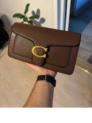 Brun kuvertväska från Coach - Snygg brun kuvertväska från Coach i slätt läder med guldfärgat metallspänne framtill. Väskan har en stilren och klassisk design med markerade sömmar och rektangulär form. Perfekt accessoar för att lyfta din outfit.