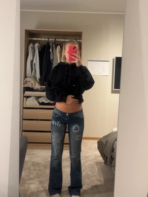 Blåa bootcut jeans med tryck - Säljer ett par blåa bootcut jeans med coola vita tryck och broderier både fram och bak. Jeansen har låg midja och slitningar på låret samt detaljerade motiv på bakfickorna. Perfekta för dig som vill sticka ut med din stil. Finns slitningar längst ner samt att knappen är lite lös men inget som märks av. Köpta för 500kr, kan tänka mig sänka priset. Midjemått: ca 42cm, innerbenslängd: 82cm