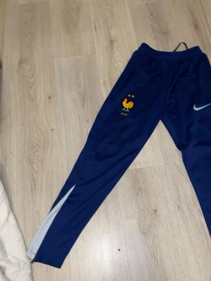 Blå Nike träningsbyxor Frankrike - Säljer ett par blå träningsbyxor från Nike med Frankrikes landslagslogga och två stjärnor broderade på benet. Byxorna har elastisk midja och en vit detalj längs benet. Perfekta för fotbollsträning eller chill.