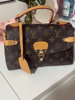  Monogram handväska - Säljer en klassisk handväska i brunt monogrammönster med detaljer i ljust läder. Väskan har guldfärgade metalldetaljer, ett elegant spänne framtill och ett topphandtag. Ikonisk design som verkligen sticker ut.