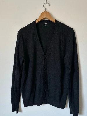 Mörkgrå kofta från Uniqlo - En stilren mörkgrå kofta från Uniqlo i 100% ull med v-ringning och knäppning framtill. Långärmad och tillverkad i ett tunt, mjukt material som passar perfekt för lager-på-lager. Enkel design som funkar till det mesta. 8D19