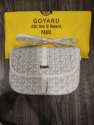 Vit axelväska  - Snygg vit axelväska med klassiskt mönster och silverfärgade detaljer. Väskan har justerbar axelrem och stängs med lock. Tillverkad i canvas med läderdetaljer. Perfekt för dig som vill ha en unik och trendig accessoar.