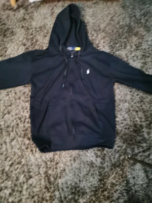 Svart hoodie från Polo Ralph Lauren - Svart hoodie från Polo Ralph Lauren med klassisk vit logga på bröstet. Tröjan har huva med snörning, dragkedja framtill och två fickor. Tillverkad i mjuk bomull, perfekt för chill dagar. Snygg och enkel design som funkar året runt.
