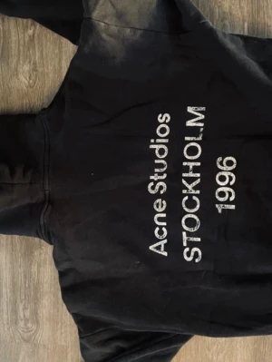 Svart hoodie Acne Studios Stockholm - Säljer en svart hoodie från Acne Studios med trycket 'Acne Studios STOCKHOLM 1996' i vitt på framsidan. Klassisk passform med huva och ribbade muddar. Perfekt för dig som gillar stilrena och trendiga plagg med streetwear-vibe.