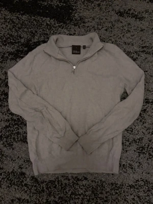 Beige quarterzip  - Säljer en stilren grå stickad tröja från Oscar Jacobsson med halv dragkedja vid kragen. Tröjan har lång ärm och ribbade muddar vid ärmslut och nederkant. Perfekt för lager på lager och enkel att matcha med olika outfits.