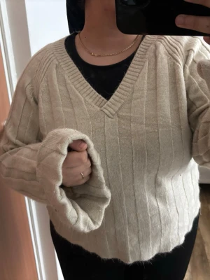 Beige ribbstickad v-ringad tröja - Mysig beige stickad tröja med v-ringning och breda ribbade ränder. Tröjan har långa ärmar och en avslappnad passform, perfekt för lager på lager. Passar till både jeans och kjol för en chill look.