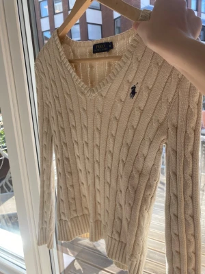Beige kabelstickad tröja Polo Ralph Lauren - Kabelstickad beige tröja från Polo Ralph Lauren med V-ringning och broderad marinblå logga på bröstet. Långärmad modell i mjukt material, perfekt för lager-på-lager och klassisk stil.