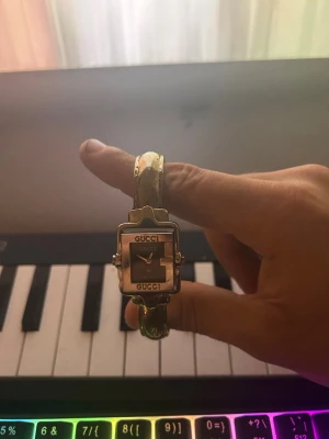 Silver vintage klocka från Gucci - Säljer en elegant klocka från Gucci med fyrkantigt ur och silver armband. Klockan har en stilren svart urtavla med Gucci-logga och ett unikt G-mönster på baksidan. Perfekt för dig som vill ha en lyxig accessoar med ikonisk design.