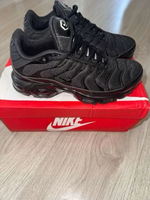 Nike Air Max Plus helsvarta sneakers - Säljer ett par helsvarta Nike Air Max Plus sneakers med mesh- och syntetmaterial, ikoniska vågiga detaljer och synlig Air-dämpning i sulan. Skorna har klassisk snörning och en sportig, chunky look som passar perfekt till streetwear.