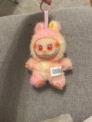 Söt fluffig monster-nyckelring rosa - Unik nyckelring i form av ett gulligt monster med fluffig päls i rosa, gult och vitt. Ansiktet är plastigt med stora ögon och ett brett leende med tänder. Har kaninöron och en BOO-lapp på magen. Perfekt att hänga på väskan eller nycklarna.