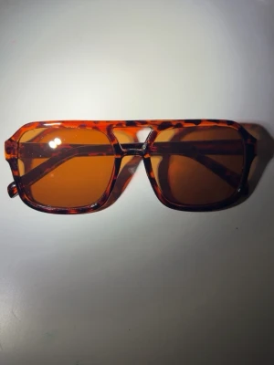 Bruna solglasögon med leopardmönster - Säljer ett par trendiga solglasögon med fyrkantiga bågar i brun plast och leopardmönster. Glasen är tonade i orangebrunt och bågarna har en cool retrostil. Perfekta för dig som vill sticka ut med en unik accessoar.