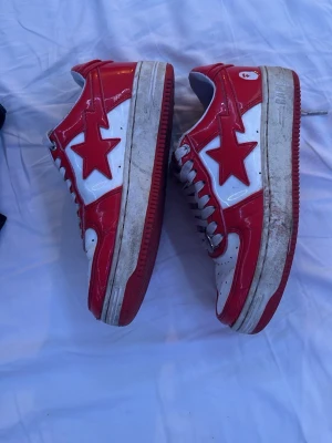 Rödvita Bape Sta sneakers - Säljer ett par ikoniska Bape Sta sneakers i rött och vitt med glansig finish. Skorna har den klassiska stjärnloggan på sidan, röda yttersulor med stjärnmönster undertill och vita skosnören. Perfekta för dig som vill sticka ut med streetwear-stil.