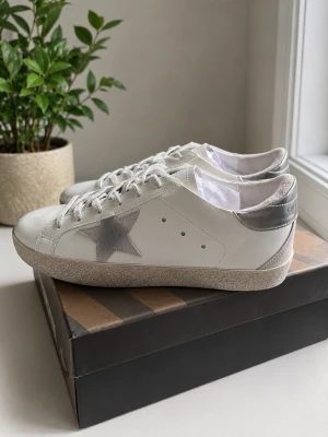 Golden Goose vita sneakers med stjärna - Snygga vita sneakers från Golden Goose med grå stjärndetalj på sidan och silverfärgad häl. Klassisk låg modell med vita skosnören och diskreta håldetaljer. Perfekta för en trendig och avslappnad stil. Påse tillkommer och skorna sitter mer som en 42.