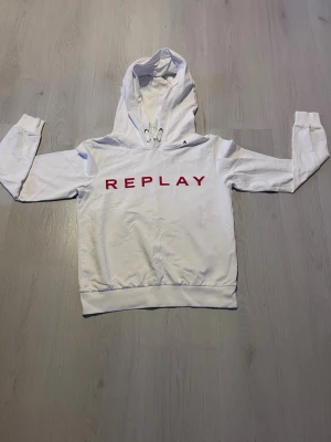 Vit hoodie från Replay med tryck - Snygg vit hoodie från Replay med röd text på bröstet. Klassisk passform med huva och dragsko, samt ribbade muddar vid ärmar och midja. Tillverkad i mjukt material som känns skönt mot huden. Perfekt för dig som gillar stilrena och trendiga plagg. Pris 400 men kan diskuteras
