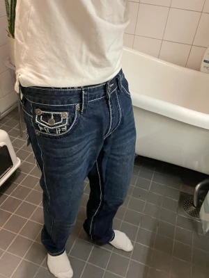 True Religion mörkblå jeans - Säljer ett par mörkblå jeans från True Religion med kontrastsömmar och stora fickor med dekorativa detaljer. Klassisk femficksmodell med tydlig logga bak och coola knappar. Jeansen har en avslappnad passform och är tillverkade i denim.