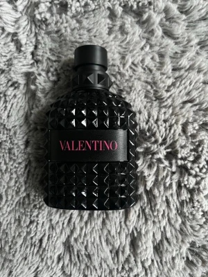 Valentino parfym svart flaska - Säljer en parfym från Valentino i en cool svart flaska med nitar och strukturmönster. Flaskan är gjord av glas och har en svart etikett med rosa text. Perfekt för dig som gillar lyxiga och snygga parfymer. Den är 100 ml men den är lite använd kanske 90 ml kvar se bild nedan 
