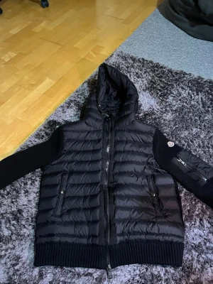 Svart dunväst med huva från Moncler - Snygg svart dunväst från Moncler med huva och stickade ärmar. Västen har två fickor med dragkedja och ribbade muddar nedtill.  Den är i storlek S men passar som xs