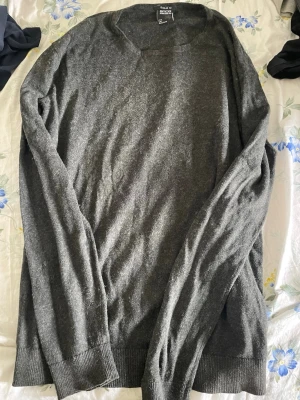 Mörkgrå stickad tröja från Smog - Mörkgrå stickad tröja från Smog i regular fit. Tröjan har rund halsringning, långa ärmar och ribbade muddar vid ärmslut och nederkant. Perfekt för lager på lager och enkel att matcha med olika stilar.