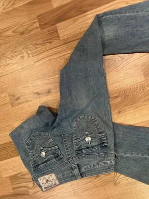 Blå True Religion jeans med fickdetaljer - Säljer ett par blå jeans från True Religion med raka ben och snygga kontrastsömmar. Baksidan har två stora fickor med dekorativa sömmar och knappar. Klassisk denimlook i 100% bomull, perfekt till en avslappnad stil. Nypris 1800kr