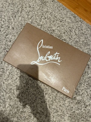 Christian Louboutin skokartong & dustbag - Säljer en original Christian Louboutin skokartong med tillhörande vit dustbag och äkthetskort. Perfekt för dig som vill förvara eller resa med dina Louboutin-skor. Dustbagen har röd logga och boxen är brun med vit text.