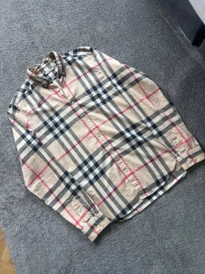 Beige rutig skjorta från Burberry - Klassisk beige skjorta från Burberry med det ikoniska rutiga mönstret i svart, vitt och rött. Skjortan har långa ärmar, knappar framtill och en bröstficka. Tillverkad i mjuk bomull och perfekt för en stilren look.