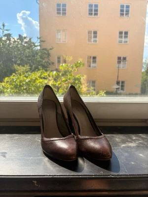 Bruna klassiska pumps i läder - Säljer ett par stilrena bruna pumps med spetsig tå och hög klack. Skorna är tillverkade i läder och har en elegant, tidlös design som passar perfekt till många olika outfits. Inga synliga detaljer eller loggor på utsidan. Perfekta för dig som gillar en klassisk look.