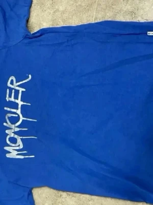 Blå Moncler t-shirt med tryck - Snygg blå t-shirt från Moncler med ett vitt grafiskt tryck på framsidan. T-shirten har klassisk passform och är tillverkad i mjuk bomull som känns skön mot huden. Perfekt för dig som gillar streetwear och vill sticka ut lite extra.