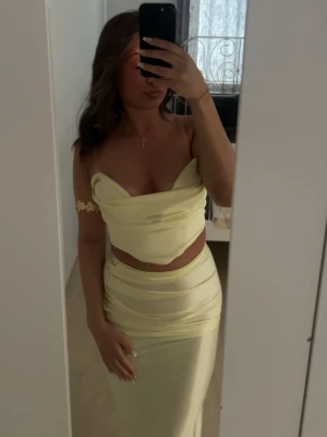 Ljusgult satinsätt med crop top - Säljer ett ljusgult tvådelat set i satin med en axelbandslös crop top och matchande lång kjol med hög midja. Materialet är glansigt och följsamt, vilket ger en lyxig känsla. Perfekt för dig som vill sticka ut med en fräsch och trendig look.