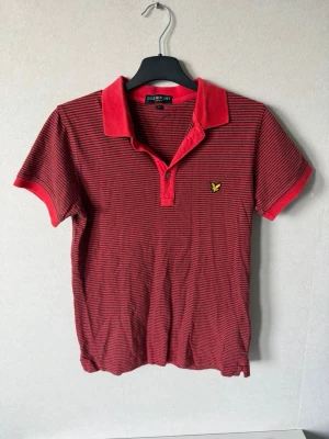 Röd randig pikétröja från Lyle & Scott - Snygg röd pikétröja från Lyle & Scott med tunna svarta ränder och klassisk krage. Tröjan har korta ärmar, knappslå framtill och den ikoniska gula loggan på bröstet. Perfekt till jeans eller shorts för en avslappnad look.