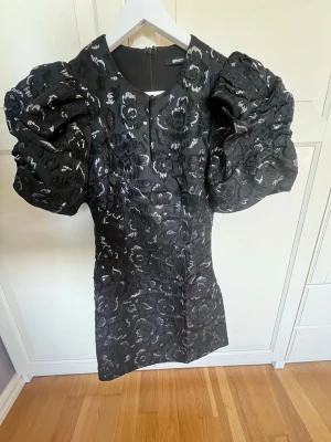Svart blommig klänning Gina Tricot - Supersnygg svart klänning från Gina Tricot med puffiga ärmar och blommigt mönster i glansigt tyg. Klänningen har en figurnära passform och dragkedja bak. Perfekt statement piece med coola detaljer och struktur. Använd en gång 