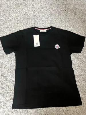 Svart t-shirt - Snygg svart t-shirt med klassisk rund hals och korta ärmar. På bröstet sitter Monclers ikoniska logga i vitt och rött. Tillverkad i mjuk bomull för en bekväm känsla. Perfekt till jeans eller shorts.