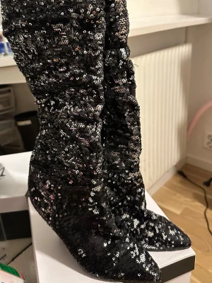 Svarta paljettstövletter med klack - Glittriga svarta stövletter täckta med paljetter och spetsig tå. Skorna har hög klack och scrunchad skaftdesign som ger en cool och trendig look. Perfekta för dig som vill sticka ut och älskar att glänsa.