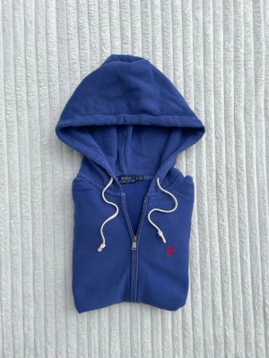 Blå zip hoodie från Polo Ralph Lauren - Säljer en blå hoodie från Polo Ralph Lauren med dragkedja, vit dragsko i huvan och klassiska röda loggan broderad på bröstet. Hoodien har kängurufickor framtill och är gjord i mjukt bomullsmaterial. Perfekt för en chill och avslappnad stil.
