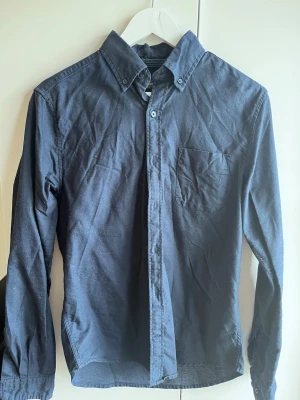 Mörkblå skjorta Dressmann slim fit - Snygg mörkblå skjorta från Dressmann i slim fit-modell. Skjortan har klassisk krage med knappar, bröstficka och långa ärmar. Tillverkad i mjuk bomull som känns skön mot huden. Perfekt för dig som gillar en stilren och enkel look.