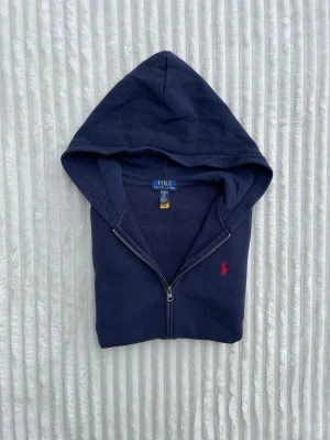 Mörkblå hoodie från Polo Ralph Lauren - Mörkblå hoodie med dragkedja från Polo Ralph Lauren. Klassisk design med huva, känguruficka och röd broderad logga på bröstet. Tillverkad i mjuk bomullsblandning som känns skön mot huden. Perfekt för en avslappnad och stilren look.