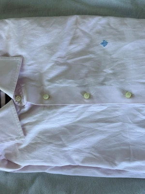 Ljusrosa skjorta från Morris - Säljer en ljusrosa skjorta från Morris med klassisk krage och knappar framtill. Skjortan har en liten blå broderad detalj på bröstet och är tillverkad i mjuk bomull. Perfekt för dig som gillar stilrena plagg med subtila detaljer.