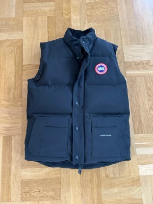 Svart dunväst från Canada Goose - Stilren svart dunväst från Canada Goose med hög krage, två stora fickor framtill och klassisk logotyp på bröstet. Västen har tryckknappar och dragkedja samt innerficka. Perfekt för lager-på-lager och håller dig varm under kyliga dagar.