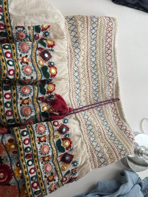 Zara kjol - Unik boho-inspirerad kjol i beige bomull med färgglada broderier i rött, blått, grönt och gult. Kjolen har dekorativa tofsar och knytband i midjan samt volangdetaljer. Perfekt för dig som gillar färg och mönster!