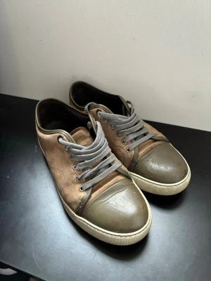 Bruna Lanvin sneakers med grå detaljer - Snygga sneakers från Lanvin i brunt läder med grå tåparti och matchande grå skosnören. Yttersulan är vit med Lanvin-logga undertill. Klassisk låg modell som passar till många olika stilar.