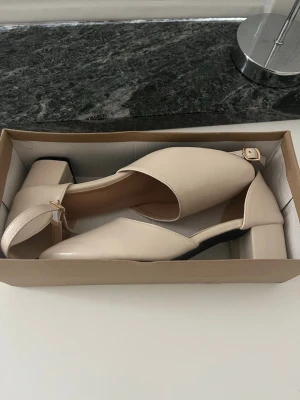 Snygga klackar - Snygga beigea klackar med låg, bred klack och rundad tå. Skorna har en elegant rem runt ankeln med spänne och är tillverkade i ett slätt, matt material som ger en stilren look. Perfekta för dig som vill ha både komfort och stil.