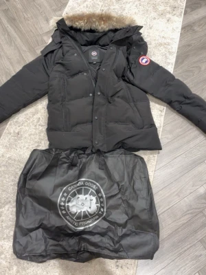 Svart dunjacka från Canada Goose - Säljer en svart dunjacka från Canada Goose med avtagbar huva och äkta pälsdetalj. Jackan har klassisk pufferstil, dragkedja och knappar framtill samt logotyp på ärmen. Perfekt för kalla vinterdagar och riktigt snygg streetstil.