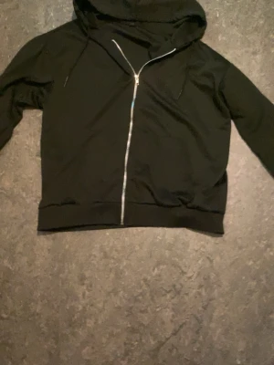 Svart zip up hoodie med huva - En klassisk svart hoodie med dragkedja framtill och huva med snören. Hoodien har långa ärmar och breda muddar vid ärmslut och nederkant. Perfekt för en chill och avslappnad stil. Materialet känns mjukt och bekvämt, troligen bomull.
