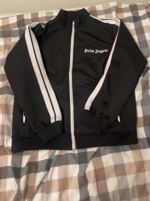 Svart track jacket från Palm Angels - Snygg svart track jacket från Palm Angels med vita ränder längs ärmarna och dragkedja framtill. Jackan har hög krage, ribbade muddar och fickor med dragkedja. Perfekt för en streetwear-look och enkel att matcha med andra plagg.