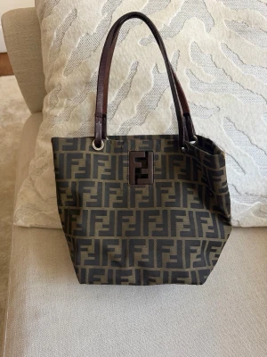 Fendi Zucca handväska brun - Säljer en klassisk Fendi Zucca handväska i brunt och beige med det ikoniska FF-mönstret. Väskan har bruna läderhandtag och detaljer samt en stor Fendi-logga framtill. Tillverkad i canvas med läderdetaljer och dragkedja på insidan.