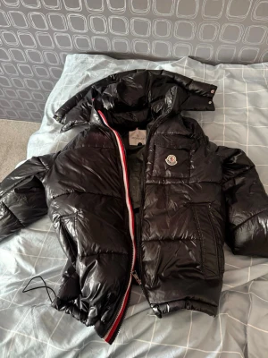 Svart dunjacka från Moncler - Snygg svart dunjacka från Moncler med glansig finish, huva och dragkedja med röd, vit och blå detalj. Jackan har en Moncler-logga på bröstet och elastiska muddar vid ärmar och midja. Perfekt för kalla vinterdagar.