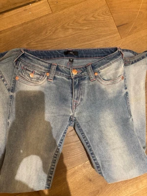 Ljusblå True Religion jeans - Säljer ett par ljusblå jeans från True Religion med klassiska kontrastsömmar i rosa. Jeansen är lite slitna längst ner. De är i W27.
