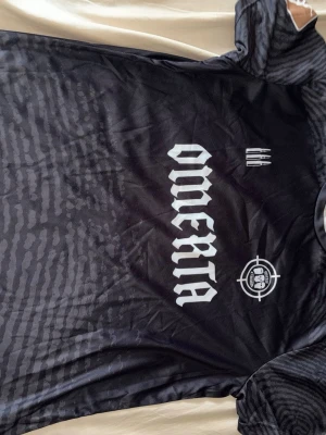 Svart Omerta t-shirt med tryck - Säljer en svart t-shirt från Omerta med stort vitt tryck på bröstet och coola grafiska detaljer. T-shirten har korta ärmar och är gjord i ett lätt, syntetiskt material som känns sportigt och skönt.