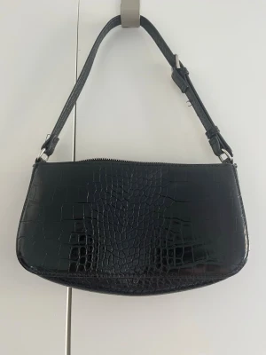 Svart axelväska i krokopräglat skinn - Snygg svart handväska med axelrem i krokopräglat konstläder. Väskan har en avlång, mjukt rundad form och stängs med dragkedja upptill. Perfekt accessoar för dig som gillar trendiga detaljer och stilren design.